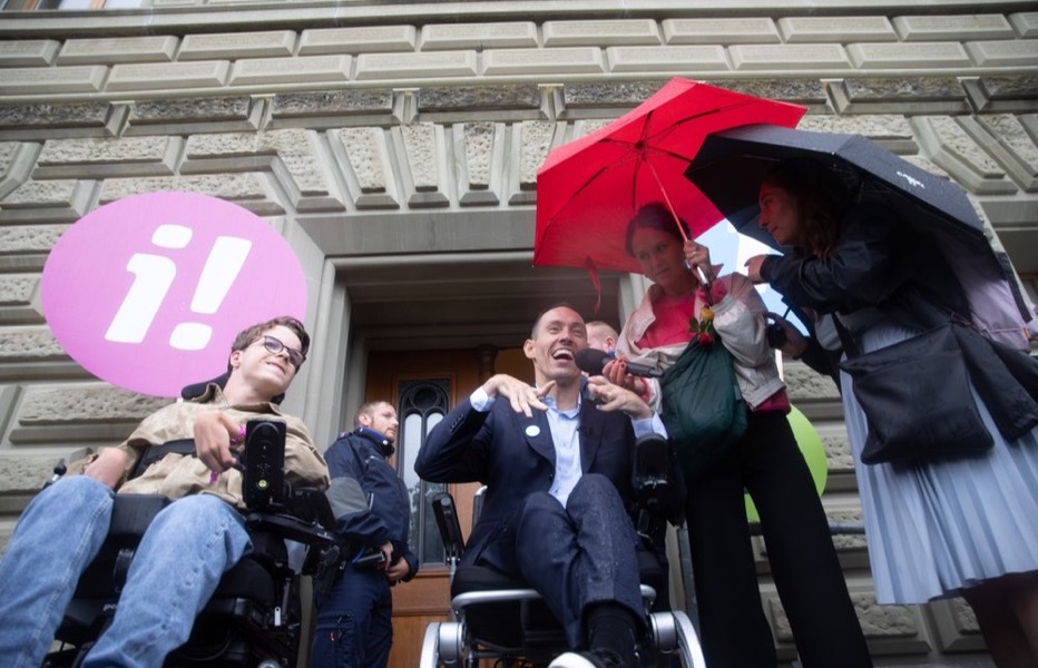Islam Alijaj an einer Kundgebung der Inklusions-Initiative vor dem Bundeshaus, im Gespräch mit Medienschaffenden unter Regenschirmen.