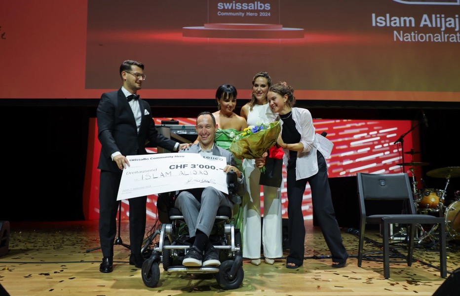 Islam Alijaj erhält auf einer Bühne den swissalbs Community Hero Award 2024, umgeben von Gratulantinnen mit Blumenstrauss.